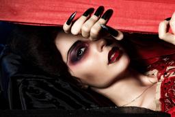 A vampire dressed in a velvety red dress peeks through a red wooden box with her long, black nails.