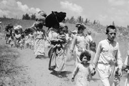 Women and children carrying bundles on their heads walk along a dirt road.