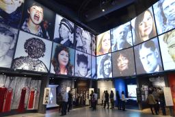 The museum's Canadian Journeys gallery, seen from the perspective of a visitor standing in the middle of it, surrounded by niches that share stories of Canadian experiences of human rights. On the walls above the niches, large images of faces of Canadian human rights defenders are seen arranged in a grid pattern.