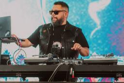 A bearded, dark-haired man, wearing sunglasses and a black t-shirt, is performing on stage using a computer and an electronic keyboard mixer.