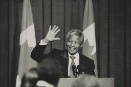 A black and white photograph of Nelson Mandela standing in front of a podium. He is smiling and waving.