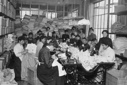 A large group of women sitting and sewing. Others are standing behind them in a room crowded with textiles. 