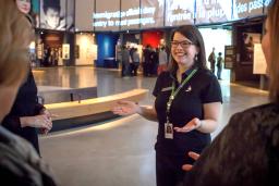 A female tour guide stands with outstretched arms in a museum gallery. Several tour participants are visible in the foreground.