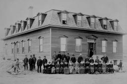 A group of Indigenous children and school officials, all wearing dark suits or dresses, pose in front of a residential school in this black-and-white photo.