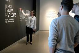 A woman points to a wall that says “Time to Act, Rohingya Voices” in both English and French while a small group of people stand and watch.