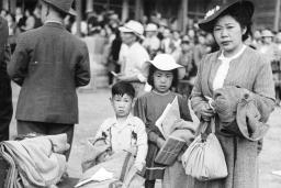 A black and white image of a woman and two children standing behind a pile of luggage and blankets and looking at the camera.