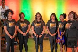 A group of six black performers standing on stage with colourful backlighting.