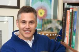 A man stands next to a bookshelf and looks into the camera.