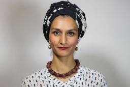 A Pakistani woman, Anam Abbas, in a black and white headwrap, red lipstick, chunky brown collar necklace and black-and-white blouse looks forward with a bemused expression.