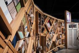 A large horizontal art installation with a wooden frame made up of multiple diamonds and rectangles to which many objects are mounted. There is a half-opened door in the middle of the work.