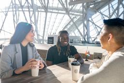 Three people sit around a table, smiling, each with a paper coffee cup in front of them. The sun shines behind them, through the panes of glass that wrap around the Canadian Museum for Human Rights.