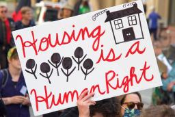 A group of people walking down a street. One person holds a sign that reads: “Housing is a human right.”