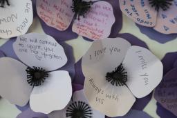 Three-petal flowers made of paper in various shades of purple are grouped together in a wall display. Names and messages are written on them. 