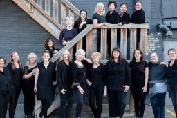 Women’s choir members are dressed in black and posed along an outdoor wooden staircase.
