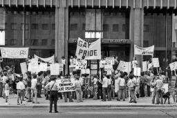A group of people protesting on the sidewalk in front of an office building. They are holding protest signs and banners and are arranged three and four deep. A police officer watches them.