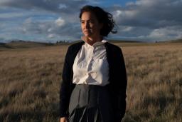 An Indigenous woman stands in a field of prairie grasses. There is a blue sky with grey clouds behind her as the sun shines on her.