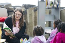 An adult sitting on the floor reading a book to three children sitting in front of them. 