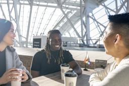Three people are seated at a table. They're all smiling with coffees in front of them. There is a small progress pride flag on their table and at the empty table behind them. The sun is shining bright through the Museum windows.