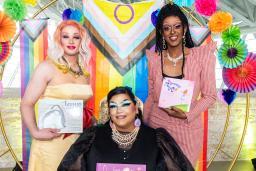 Three drag artists holding books are standing in front of a rainbow-themed circle arch.