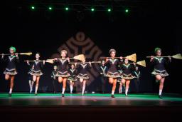 Thirteen Irish dancers dressed in short, black dresses are holding brooms across their chest.