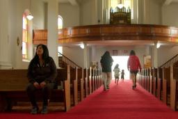 A church interior with a long red-carpeted aisle between wooden pews. Sunlight filters through stained-glass windows as several children run toward the bright entrance at the far end while an Indigenous woman sits alone in a pew to the left.