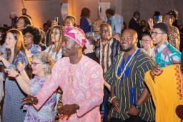 A group of people of all ages, many in brightly coloured traditional African attire, dancing and smiling.