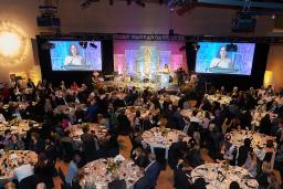 In a large hall, hundreds of diners sit at round tables while a woman speaks at a podium.