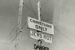 Black and white photo of three signs on a post, reading “Christians only,” “Jews not allowed” and “Danger.”