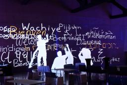 Interactive screen depicts human figures writing “welcome” in several different languages.