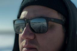 A close-up of a man wearing sunglasses and a hat, looking at something off camera. A road is seen reflected in his sunglasses.