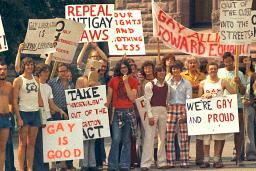 A large group of people dressed in T-shirts and seventies-style flared pants, stand together holding signs with messages such as “Out of the Closet,” “Gay is Good” and “Repeal Anti-Gay Laws.”