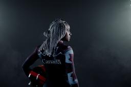 A black woman stands in a dark place between two large projectors. She's holding a helmet and wears a Canadian bobsleigh suit.