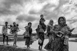 A group of Rohingya women and men carry young children and belongings as they walk in a line over an earthen dike over a stretch of water.