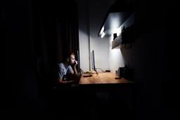 A white man with short hair sits alone at a brightly lit desk in a dark room, staring at a computer monitor.