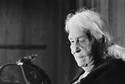 A black and white image of an elderly woman with white hair holding a mask that is shaped and painted to represent a bird’s head. It features a long beak with an open mouth and feathers attached to the top of the head. The woman is looking directly at the mask she is holding.