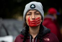 A young woman with braided hair, wearing a maroon jacket and gray winter cap, looks forward resolutely. A red handprint is painted over her mouth and face.