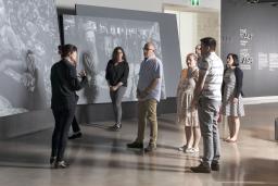 A small group of people stand in front of large screens displaying photographs and look at a woman who is speaking to them.