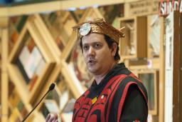 A man wearing traditional Indigenous regalia and headdress stands in front of an art installation made of wooden panels with embedded objects.