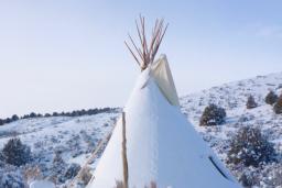 A snow-covered tipi sits in a hilly landscape dotted with trees and bushes against a blue sky.
