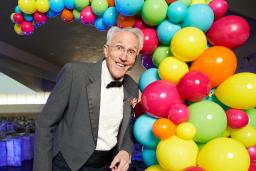 A performer wearing a tuxedo stands in front of a display of multi-coloured balloons.