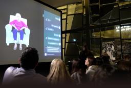 Audience members view a large projection screen displaying an elderly woman in a purple top and blue pants sitting in a white armchair, alongside an interactive message interface. The presentation takes place in a modern space with geometric glass architecture, and a presenter stands to the right of the screen.