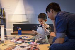 A young child interacting with photos on a table. An adult sits beside him.