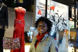 A smiling woman stands in front of a red prom dress and a black tuxedo mounted on mannequins. Both the dress and suit are on display behind a glass case.