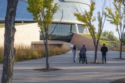 Four individuals follow the path toward the Canadian Museum for Human Rights on a fall day. Three are on foot and one is in a wheelchair.