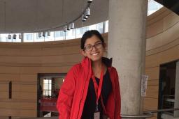 A young woman sits on a ledge in a large circular hall. She is smiling at the camera and wearing jeans, a dark blouse and a red jacket