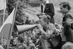 A woman wearing a dress cups her hand to a megaphone placed at her mouth. She is standing elevated above a crowd of people at a gate, with two flags waving nearby.