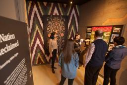 A guide speaks to a small group of people in a museum exhibit space. The space includes a wall featuring a Métis artwork and a text panel that reads “A Nation Reclaimed”.