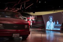 Red Chevrolet van in an exhibition space with video by Rage Against the Machine playing in the background.