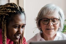 A young woman who looks to be in her teen years smiles as she works at a computer alongside an older, white-haired woman who is also smiling.