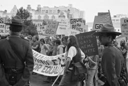 A large group of protestors march through a city while two uniformed RCMP officers keep watch. In this black-and-white image, the protestors' signs and banners call for an end to discriminatory practices against members of the 2SLGBTQI+ community.